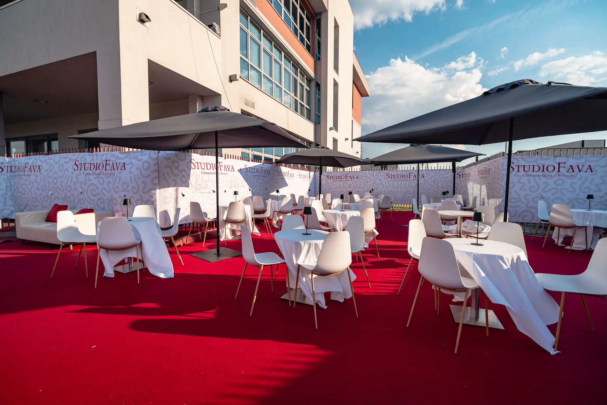 Area lounge esterna arredata da Stanport per Studio Fava, con tavoli rotondi, sedute bianche, ombrelloni e tappeto rosso